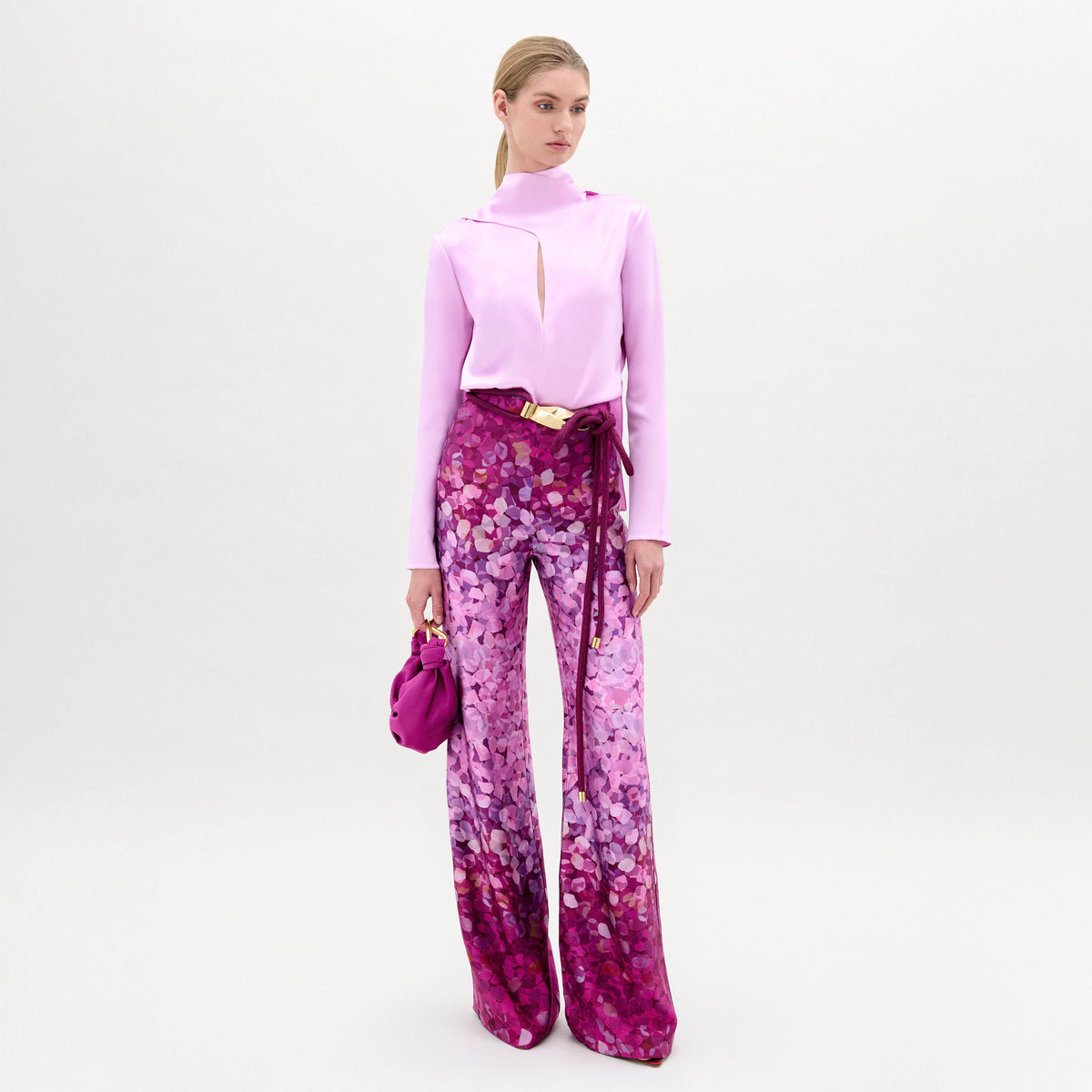 A woman stands against a white background wearing a light pink keyhole long-sleeve top, the Como Pant—high-waisted wide-leg magenta Italian floral pants—and holds a matching pink handbag.