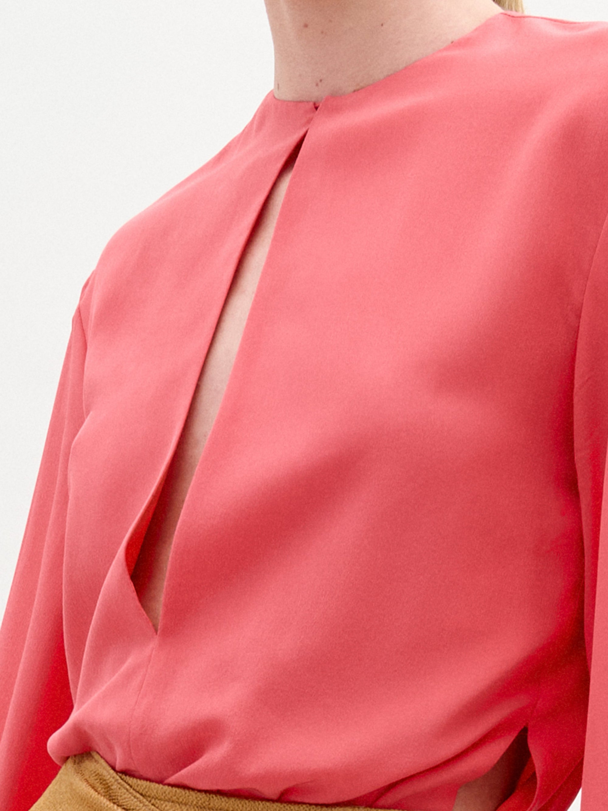 A woman stands against a plain background wearing the Covilha Blouse, a silk coral long-sleeve top, paired with a tan skirt featuring a large gold circular belt detail.
