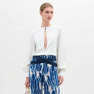 A woman stands against a plain background in the Covilha Blouse, a white silk top with a front keyhole cutout, paired with patterned blue and white high-waisted pants. She holds a small light-colored handbag, her pulled-back hair accenting the voluminous silhouette.