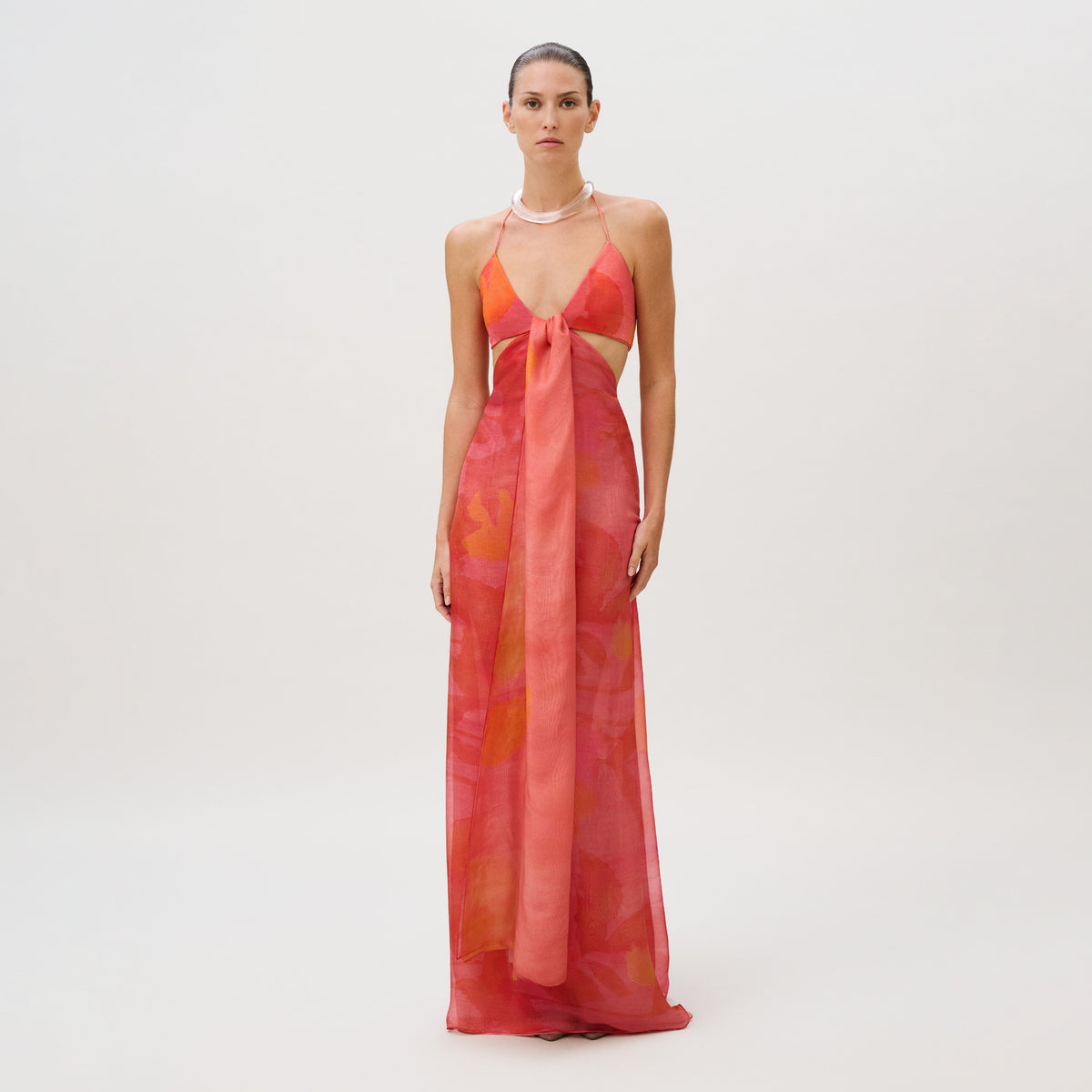 The Candida Dress is a slim-fit, floor-length, sleeveless halter dress in shades of orange and pink, crafted from jersey chiffon with a tie-front cutout detail.
