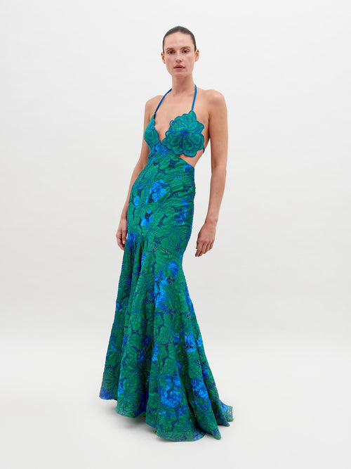 A person stands wearing a vibrant green and blue floral-patterned dress with an open back and a halter neck, posing against a plain white background. The Cara Dress Green Navy Embroidery is available for pre-order now, with a ship by date of November 15th, 2024.