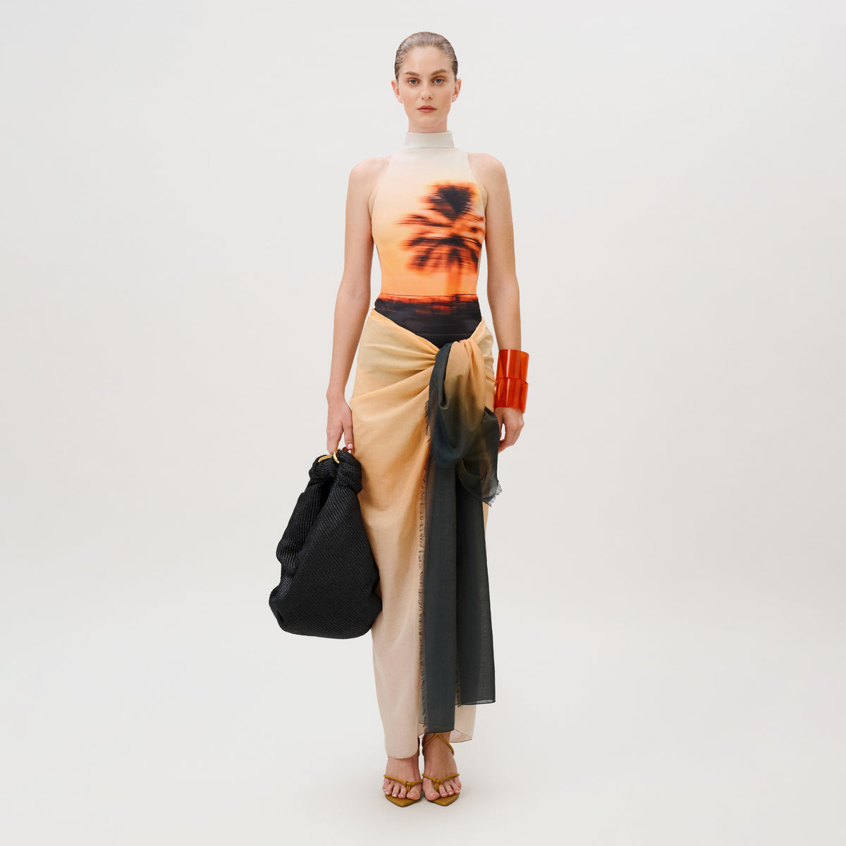 A person wearing a tropical sunset print sleeveless top, a Caribbean Pareo sheer layered Italian fabric wrap skirt, and a large black handbag.