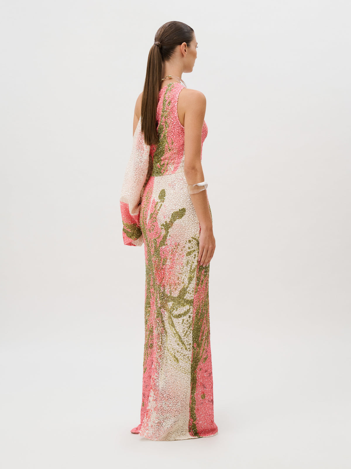 The Carmel Dress is a sleeveless, floor-length sequin gown featuring a pink and green floral pattern crafted from Italian fabric with a single draped sleeve.