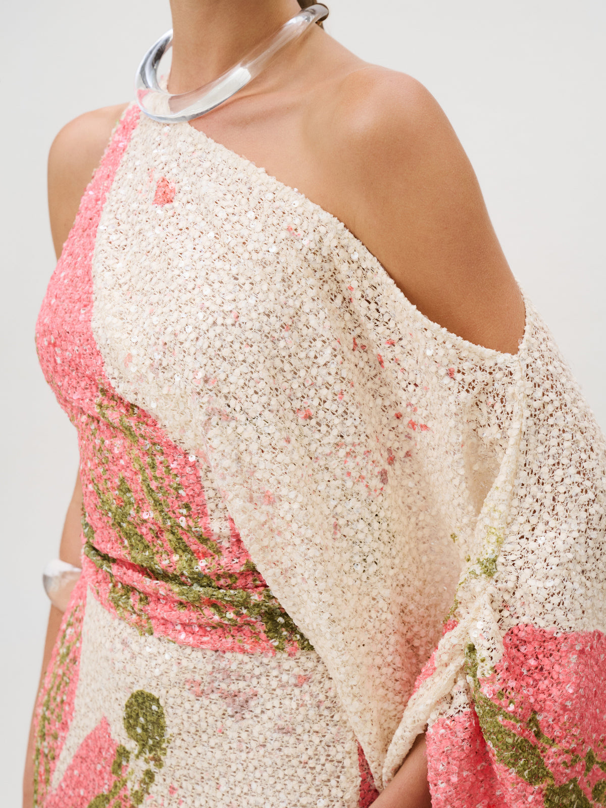 Carmel Dress, a one-shoulder dress made of open-weave Italian fabric with pink and green sequin floral patterns, worn with a clear collar necklace.