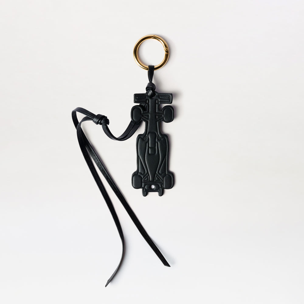 The F1 Charm is a black Formula 1 car-shaped keychain featuring a gold key ring and slim black leather straps, displayed against a white background.