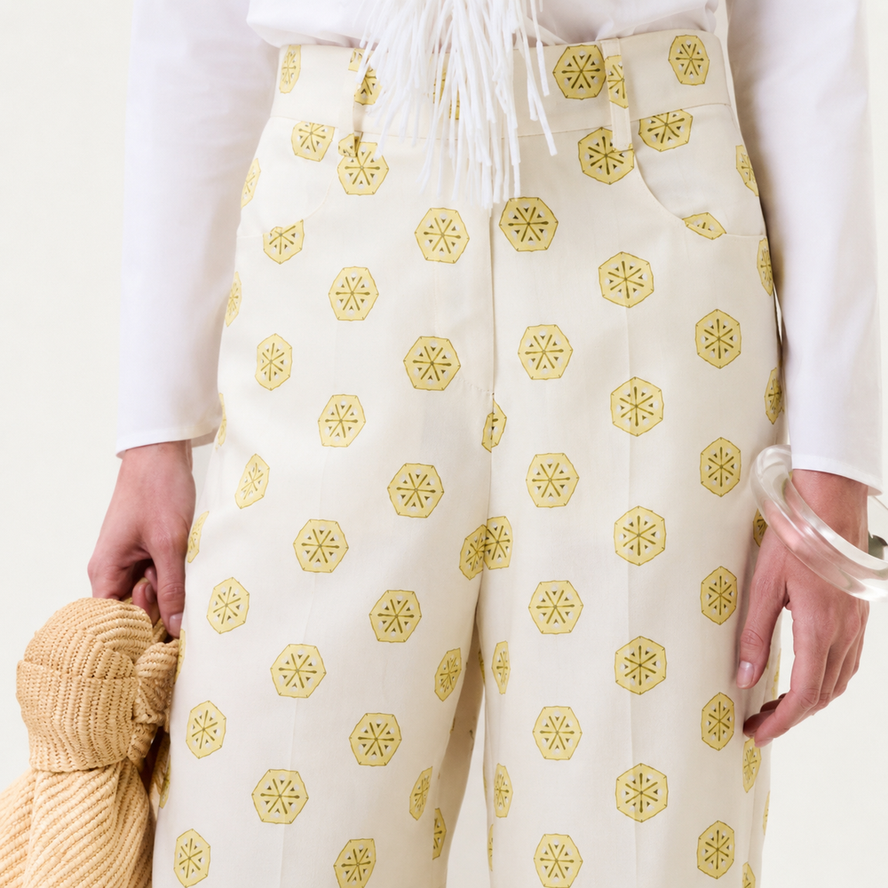A person wearing the Emine Pant—a cream pant with yellow hexagonal patterns—paired with a white top featuring white fringe detail, holding a yellow textured bag, and accessorized with a clear bangle on one wrist.
