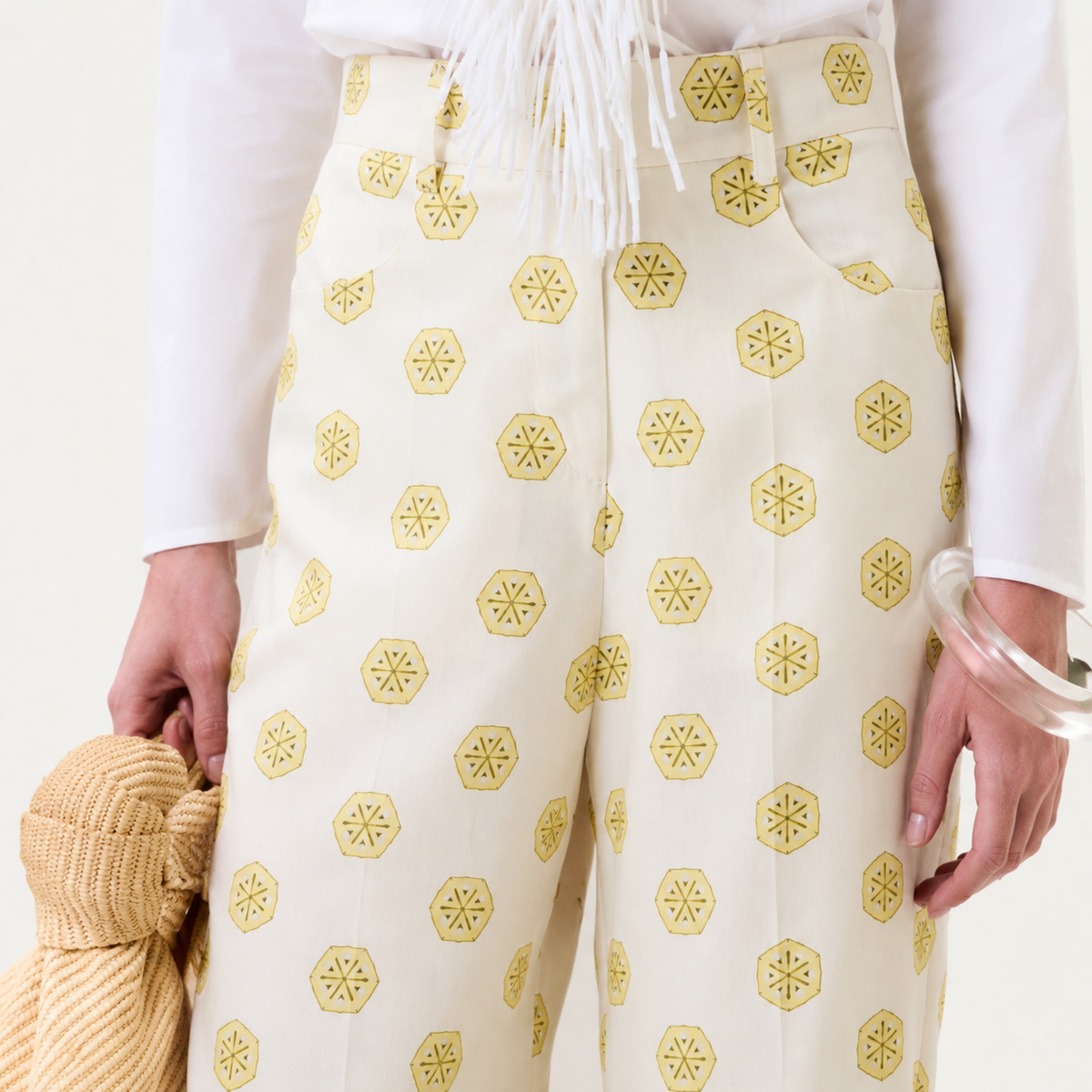 A person wearing the Emine Pant—a cream pant with yellow hexagonal patterns—paired with a white top featuring white fringe detail, holding a yellow textured bag, and accessorized with a clear bangle on one wrist.