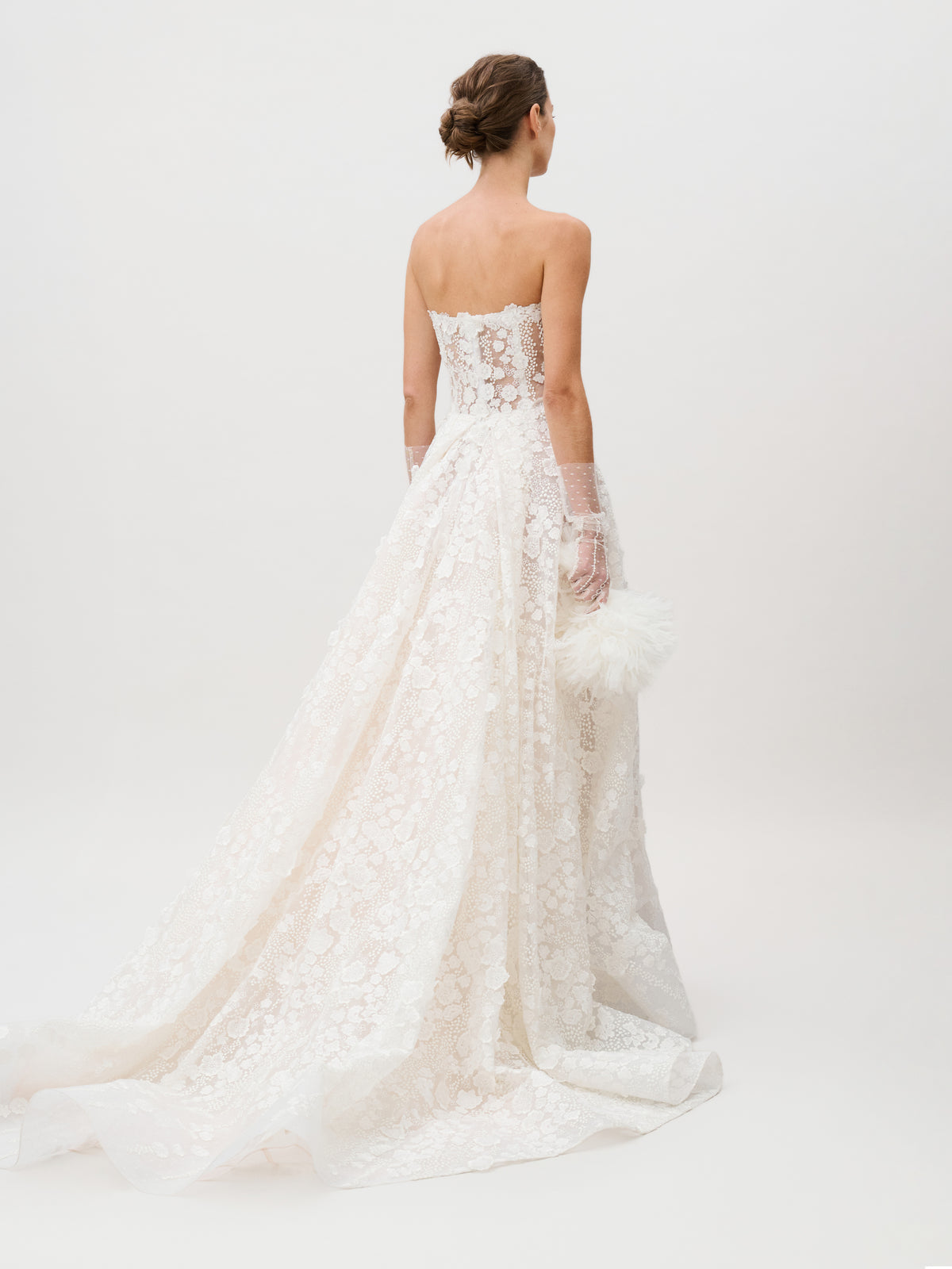 A woman faces away, holding a white, fluffy clutch while wearing the Cindy Dress—a strapless lace wedding gown expertly crafted from luxurious Italian fabric by a renowned Colombian atelier. The dress showcases a long, elegant train and sheer gloves against a plain white background.