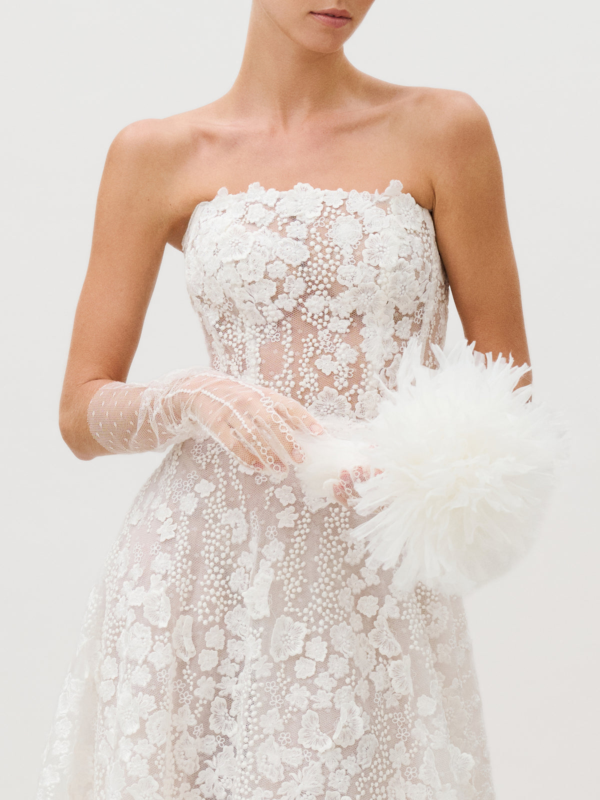 A woman wearing the Cindy Dress—a strapless white lace design featuring floral patterns—stands against a plain light background. Created by a Colombian atelier from luxurious Italian fabric, the dress is paired with sheer gloves and a fluffy white accessory. The image is cropped above her nose.
