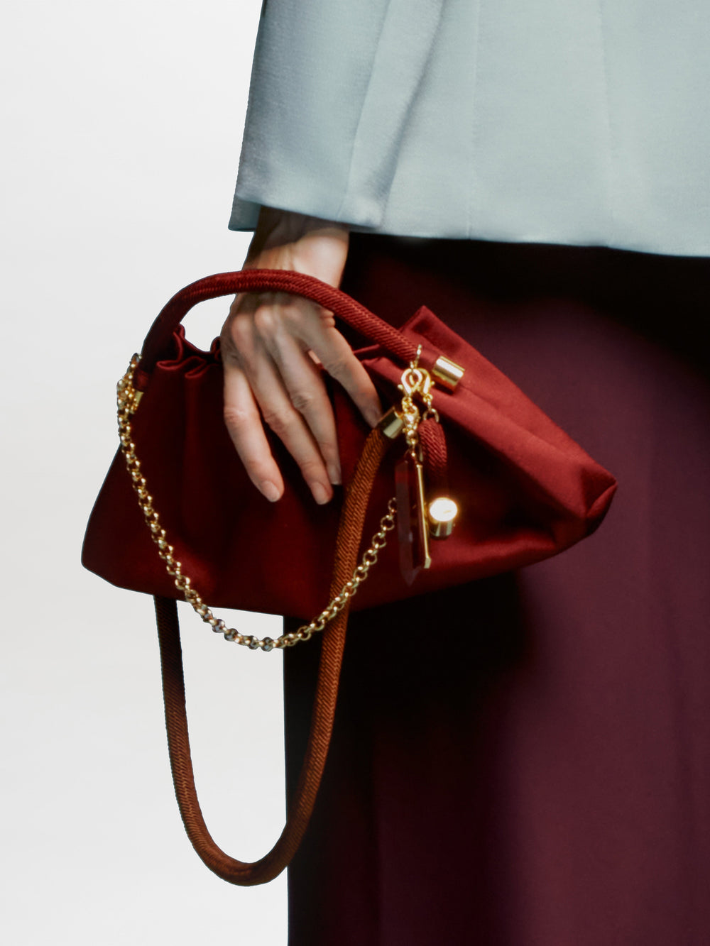 The Trapezius Clutch is a maroon pleated crepe handbag with gold accents, a textured handle, and a gold letter "S" charm.