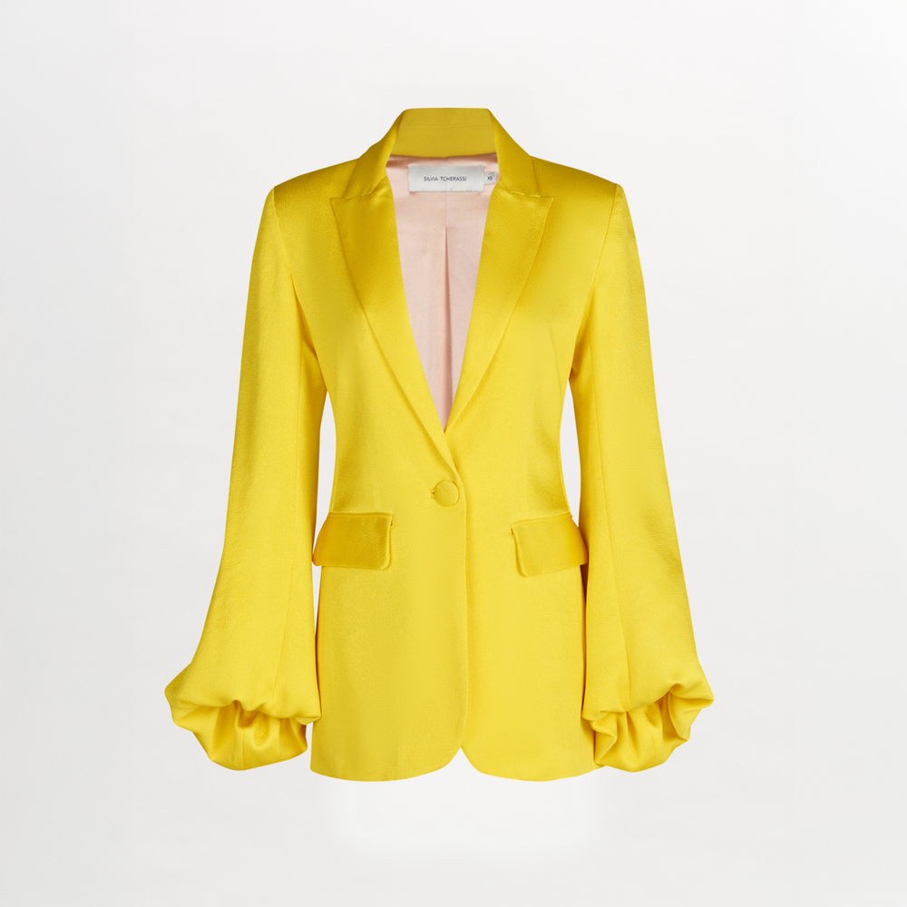 The Coco Blazer is a bright yellow hammered satin jacket with soft tailoring, a single-button closure, two front flap pockets, and voluminous gathered sleeves, shown on a plain white background.