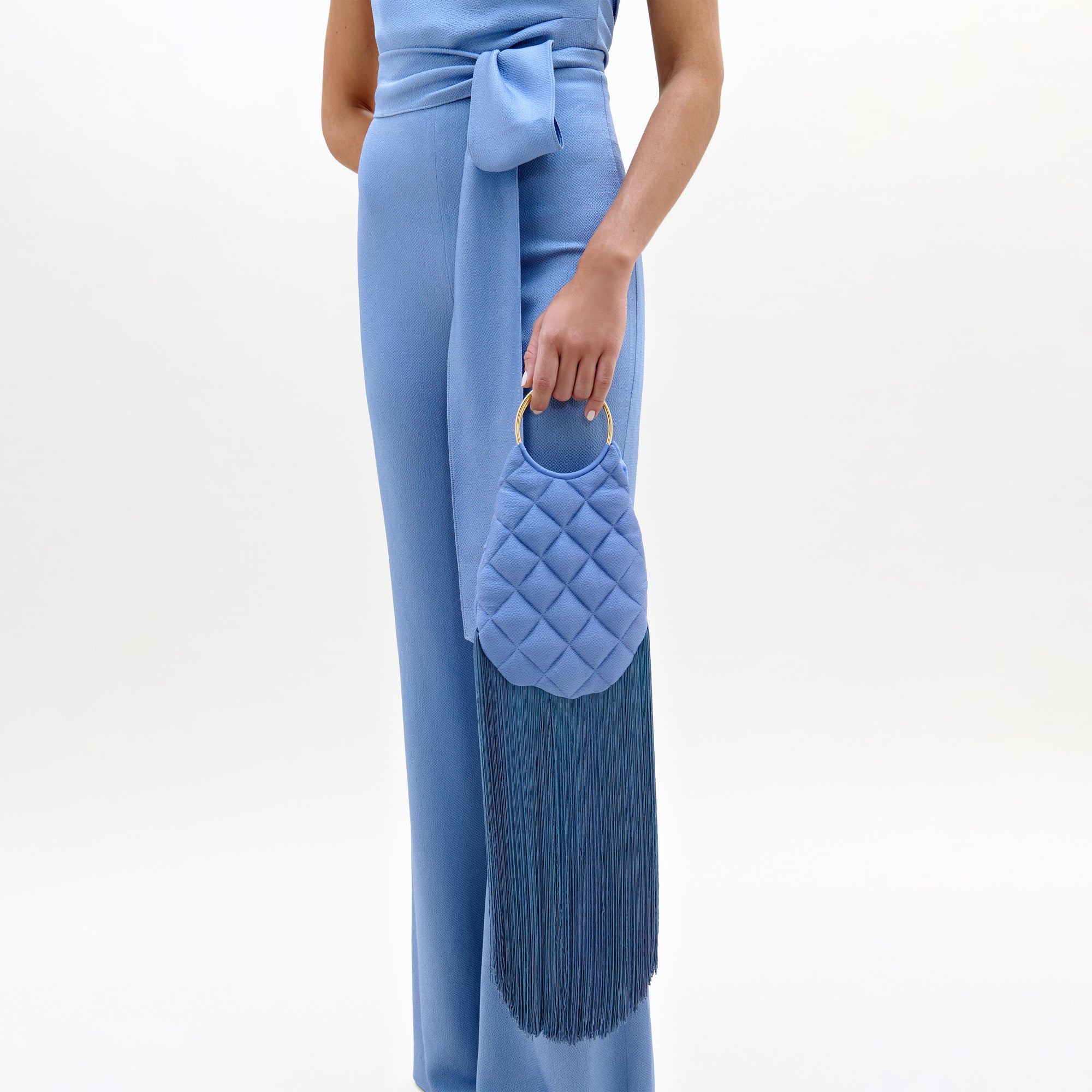 A person in a light blue jumpsuit with a sash holds the Toblach Bag, a structured oval Italian satin top-handle bag in light blue, featuring a gold ring handle and long blue fringe tassels. The background is plain white.