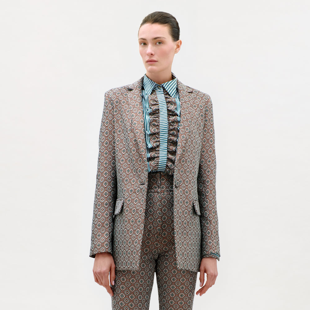 A person stands against a plain background wearing the Cuneo Jacket and matching trousers, expertly tailored from fine Italian fabric, with a striped, ruffled shirt. Their straight brown hair is pulled back, and they have a neutral expression.