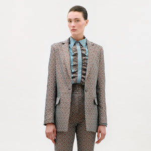 A person stands against a plain background wearing the Cuneo Jacket and matching trousers, expertly tailored from fine Italian fabric, with a striped, ruffled shirt. Their straight brown hair is pulled back, and they have a neutral expression.