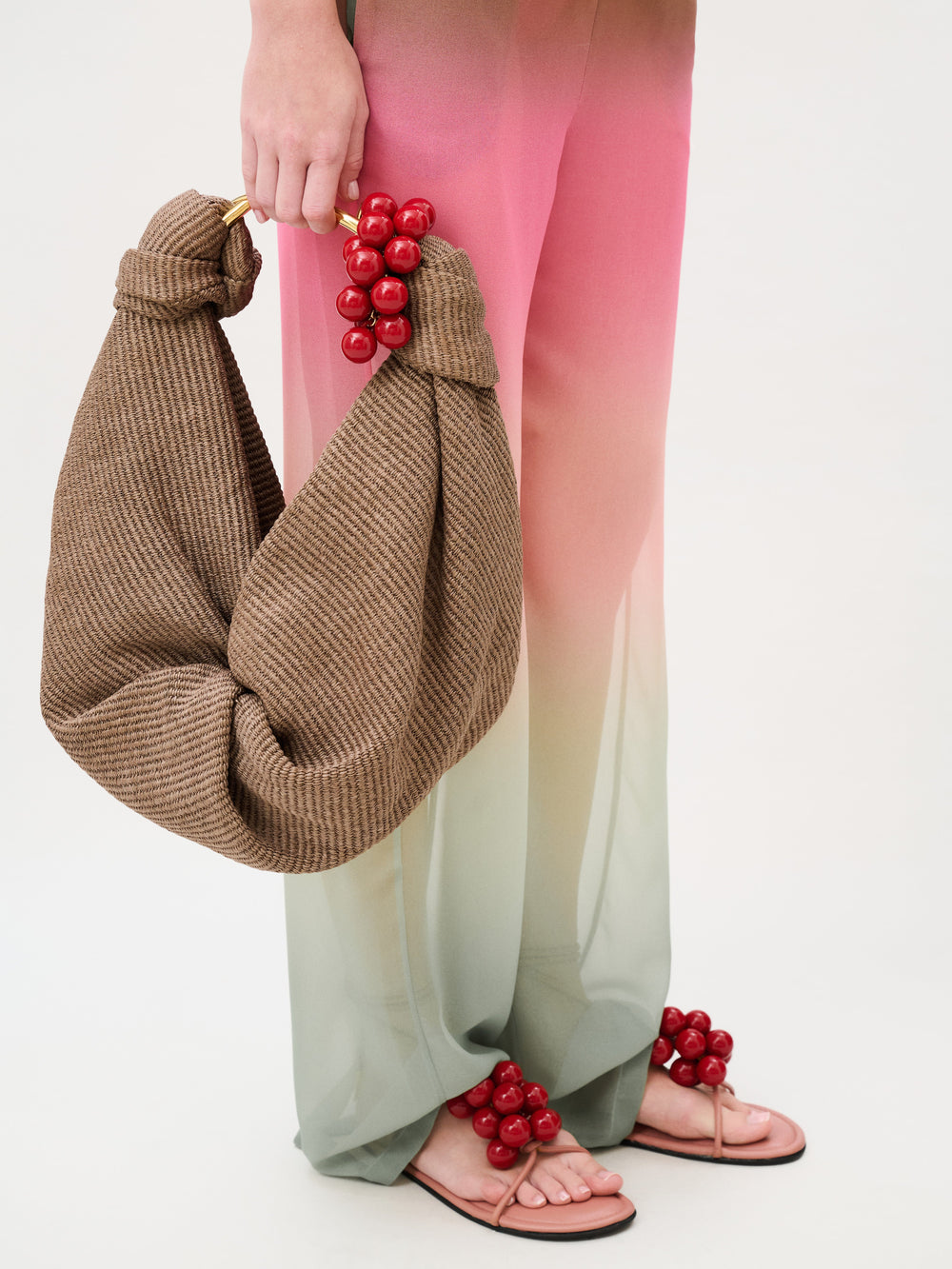 A person wearing Curacao Pant chiffon pants in an ombré transition from pink to pale green, paired with pink thong sandals adorned with red spheres, holding a brown woven hobo bag with a red bead embellishment.