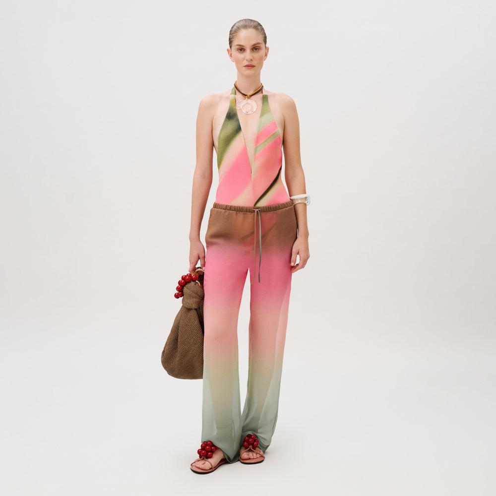 A model wears a pink and green gradient halter bodysuit and matching Curacao Pant, carrying a brown beaded tote bag and wearing matching beaded sandals.