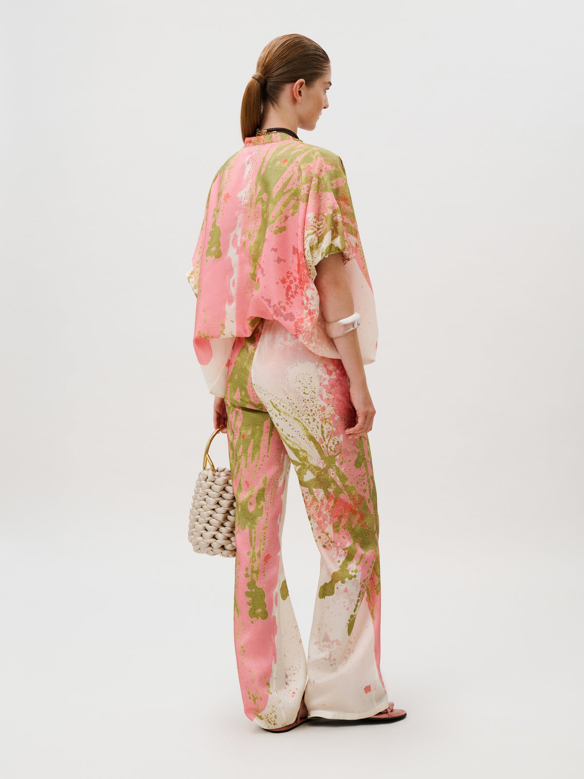 A person wears a matching loose-fitting shirt and wide-leg Curacao Pant in an abstract pink and green paint-splatter print, carrying a woven handbag.