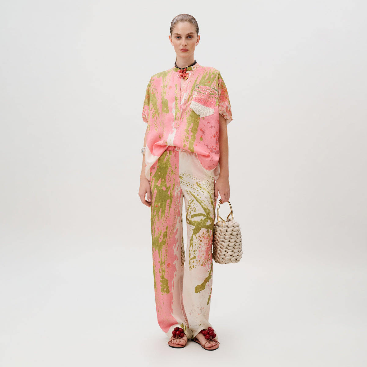 A person wearing a short-sleeved shirt and matching Curacao Pant in Italian fabric, both featuring a pink and olive green paint-splatter pattern, carrying a woven handbag.