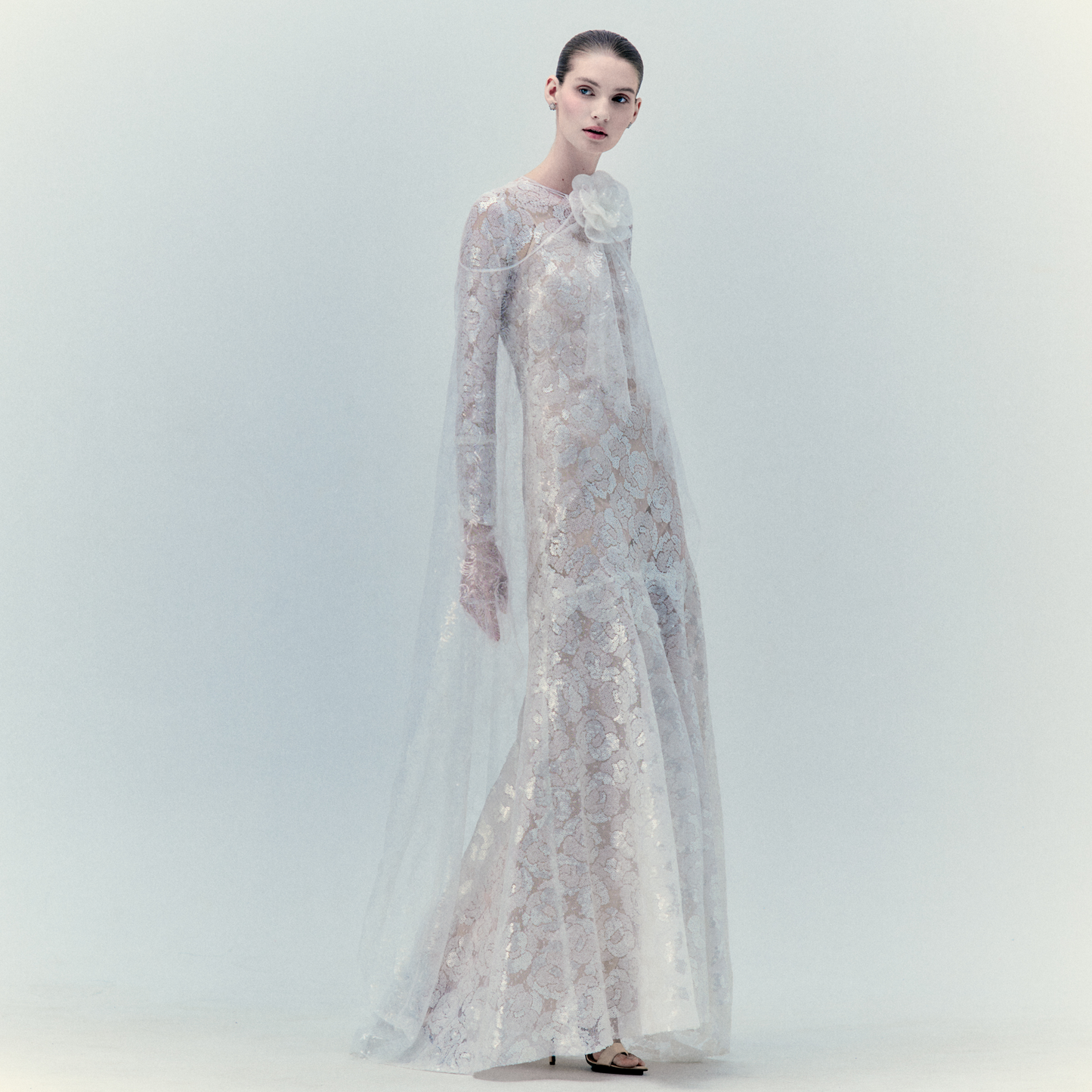 A woman poses elegantly against a light background wearing the Curie Dress, a long, sheer lace maxi featuring floral embroidery and a high neckline with a statement bow. Her hair is slicked back and her expression is soft and neutral.
