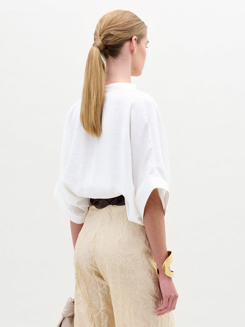 A woman wears the Daia Blouse in an oversized white fit with a V-cut neckline, paired with cream Italian fabric pants, a gold belt and accessories, and holds a small bag while posing against a white background.
