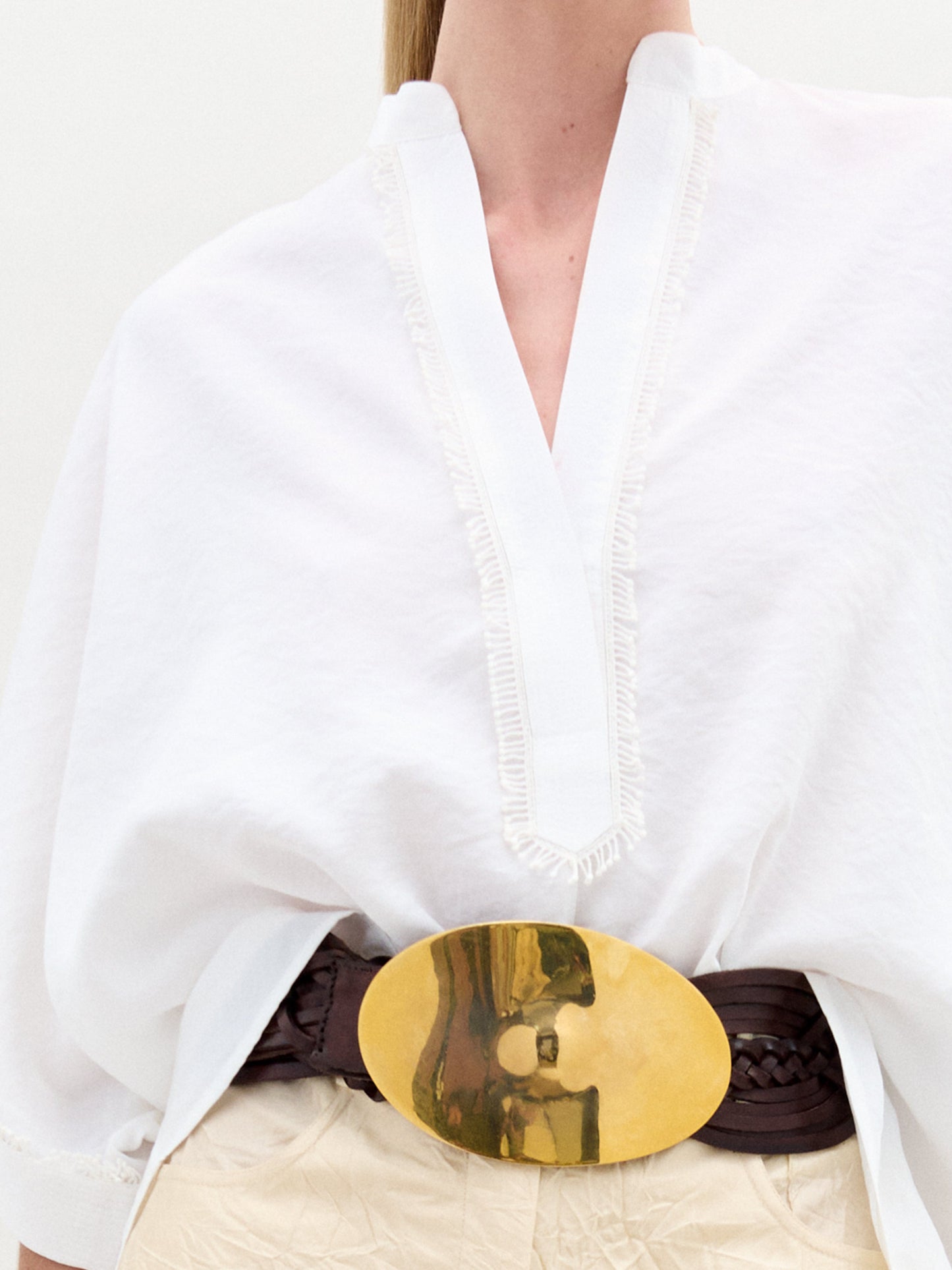 A woman wears the Daia Blouse in an oversized white fit with a V-cut neckline, paired with cream Italian fabric pants, a gold belt and accessories, and holds a small bag while posing against a white background.