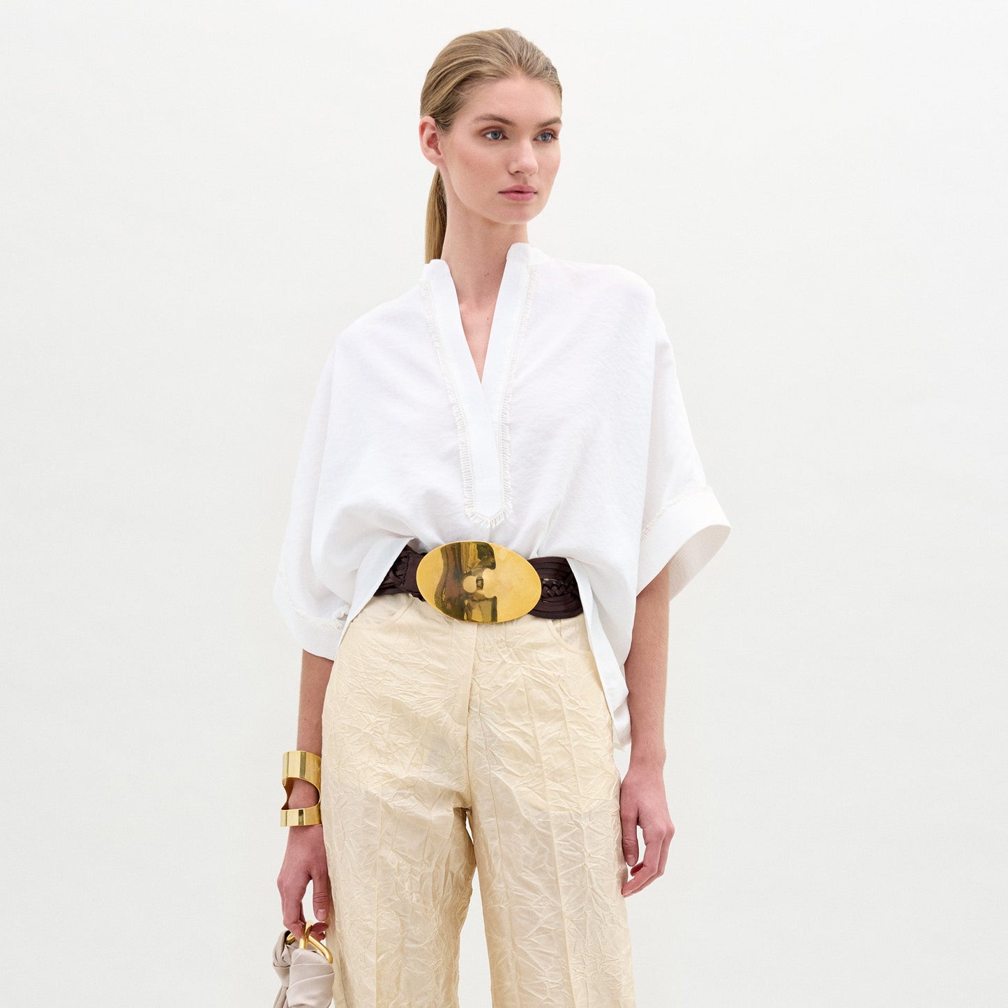 A woman wears the Daia Blouse in an oversized white fit with a V-cut neckline, paired with cream Italian fabric pants, a gold belt and accessories, and holds a small bag while posing against a white background.