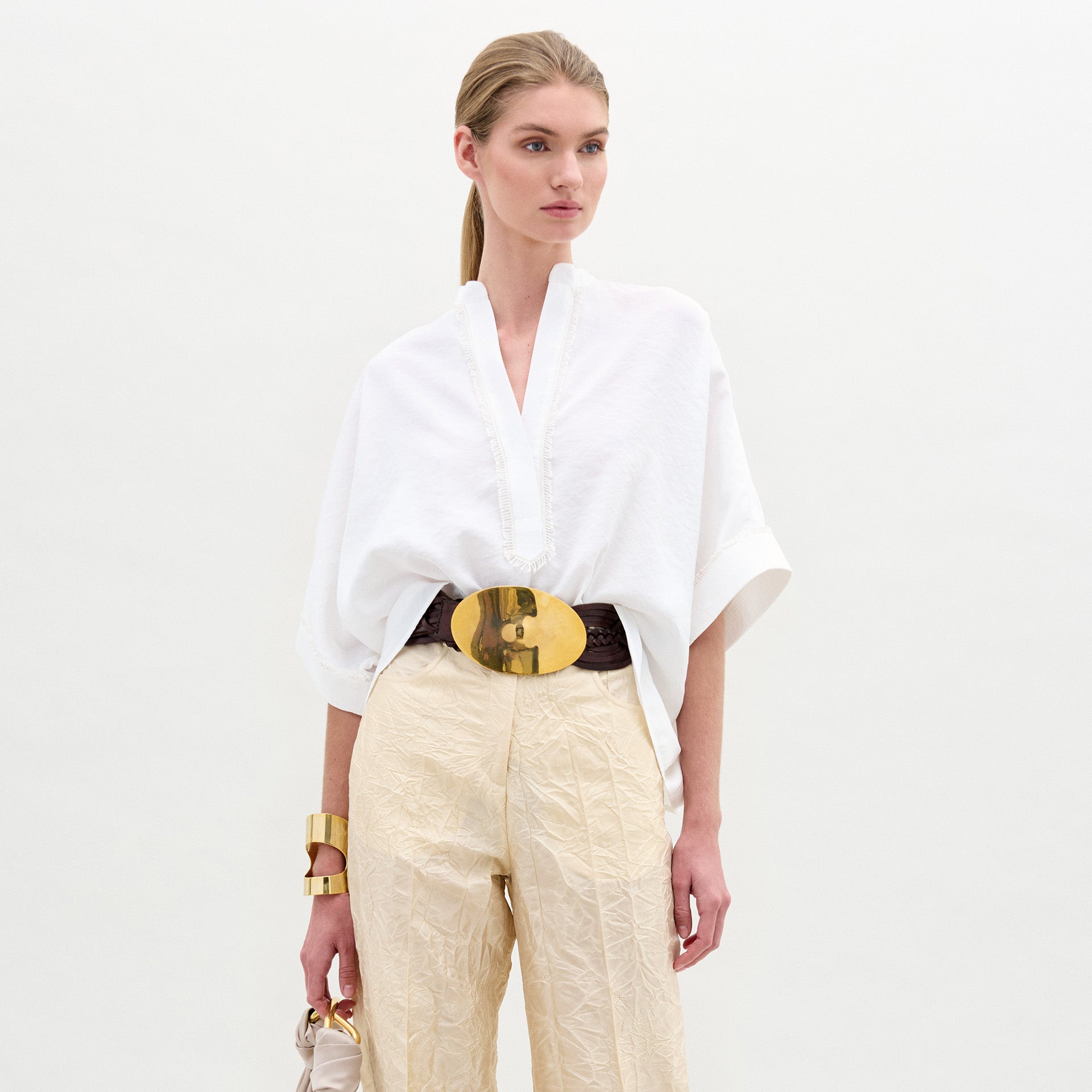 A woman wears the Daia Blouse in an oversized white fit with a V-cut neckline, paired with cream Italian fabric pants, a gold belt and accessories, and holds a small bag while posing against a white background.