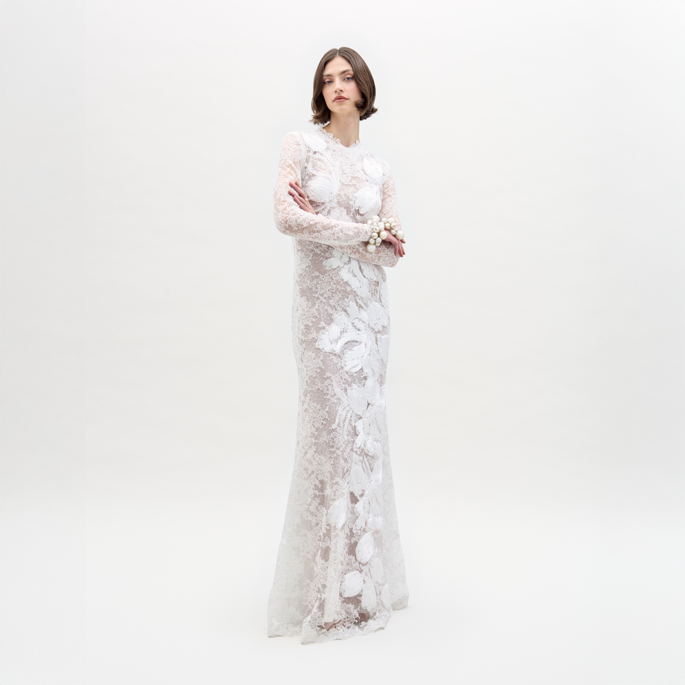 A person stands against a plain white background, arms crossed, wearing the Dava Dress—a sheer, long-sleeved lace dress with floral patterns.