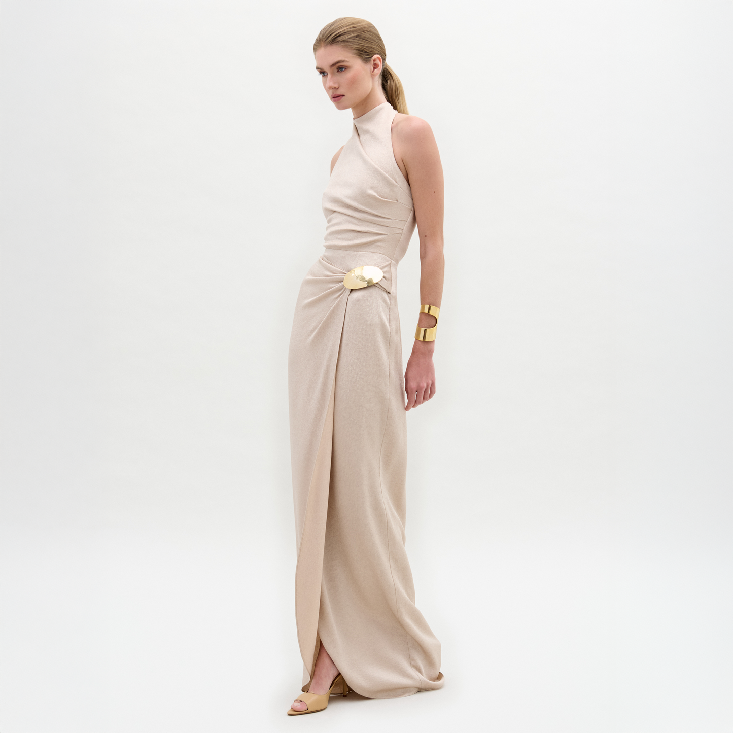 A woman wears the Davinia Dress, a cream, floor-length halter gown with a floral accent at the waist, paired with a gold cuff bracelet and beige shoes, standing against a plain white background.