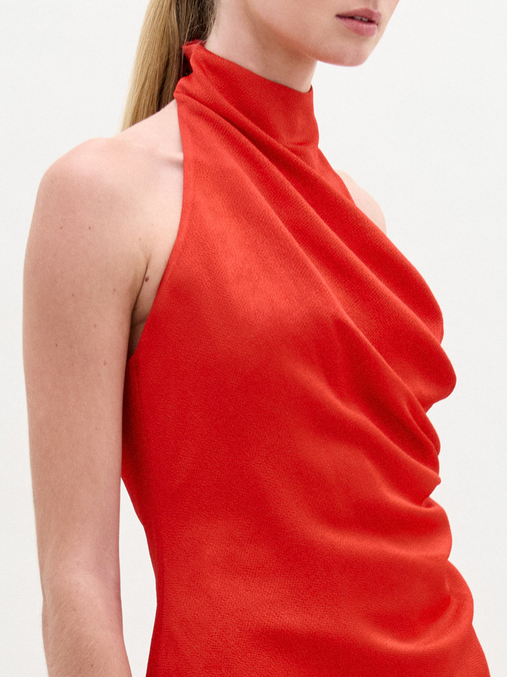 A woman models the Dina Blouse, a high-neck sleeveless top in bright red Italian fabric with draped details; her hair is pulled back and her face is partially visible against a plain background.