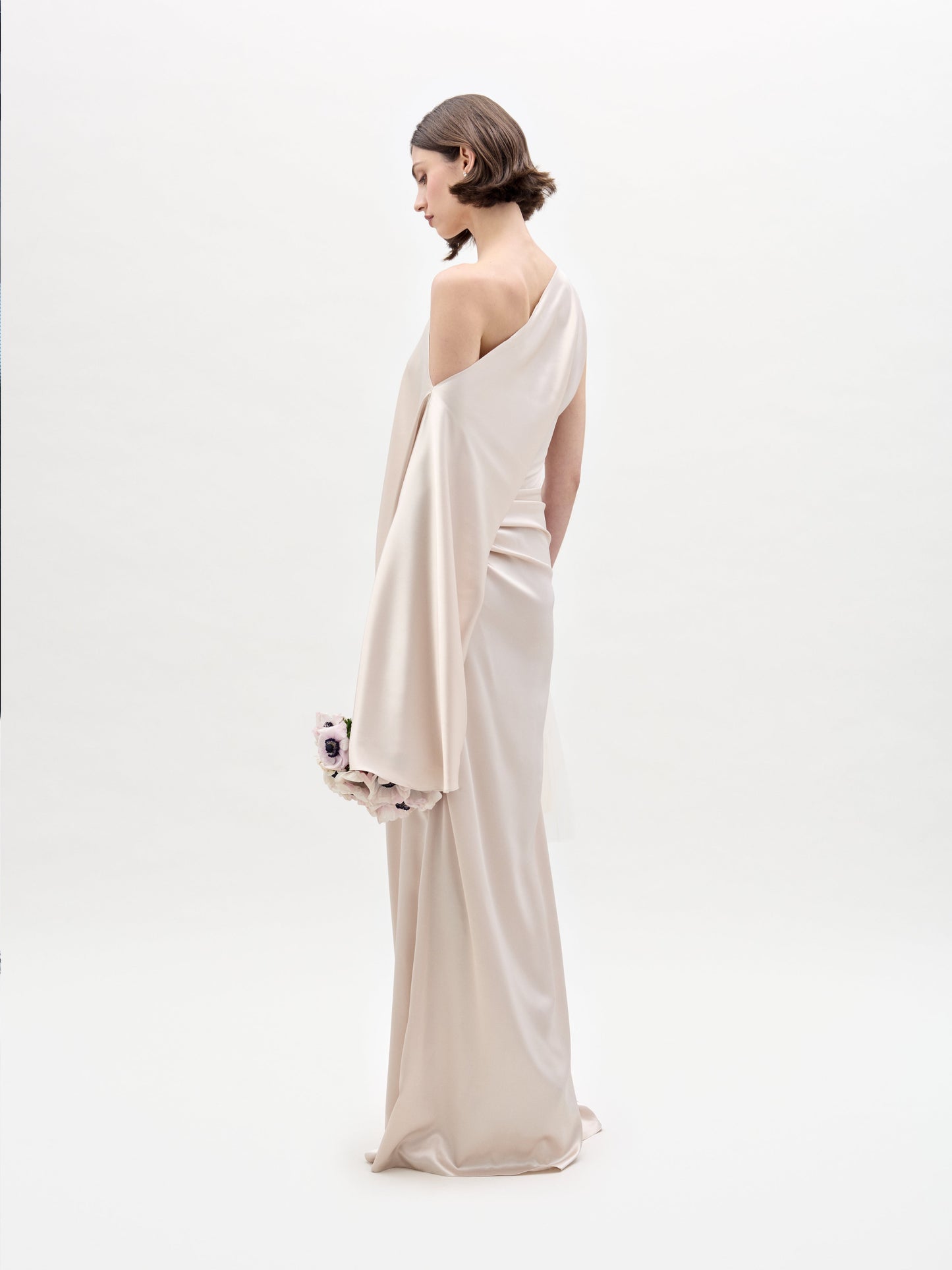 A person stands in a Dotty Dress, an off-the-shoulder satin gown, holding a bouquet of white and purple flowers against a plain white background.