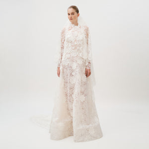 Dressed in the Danita Dress—a long, white, lace wedding gown adorned with floral patterns and crafted from exquisite Italian fabric by Colombia Atelier—a woman stands against a plain white background. Her timeless bridal look is completed with a matching lace veil.