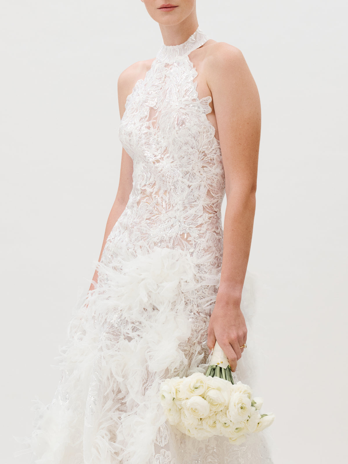Clad in the Darsha Dress—a sleeveless lace wedding gown adorned with delicate silk trim, intricate floral motifs, and ethereal feather accents—she holds a bouquet of white roses. The light background spotlights the exquisite details of this Italian-made dress, her face softly out of frame.
