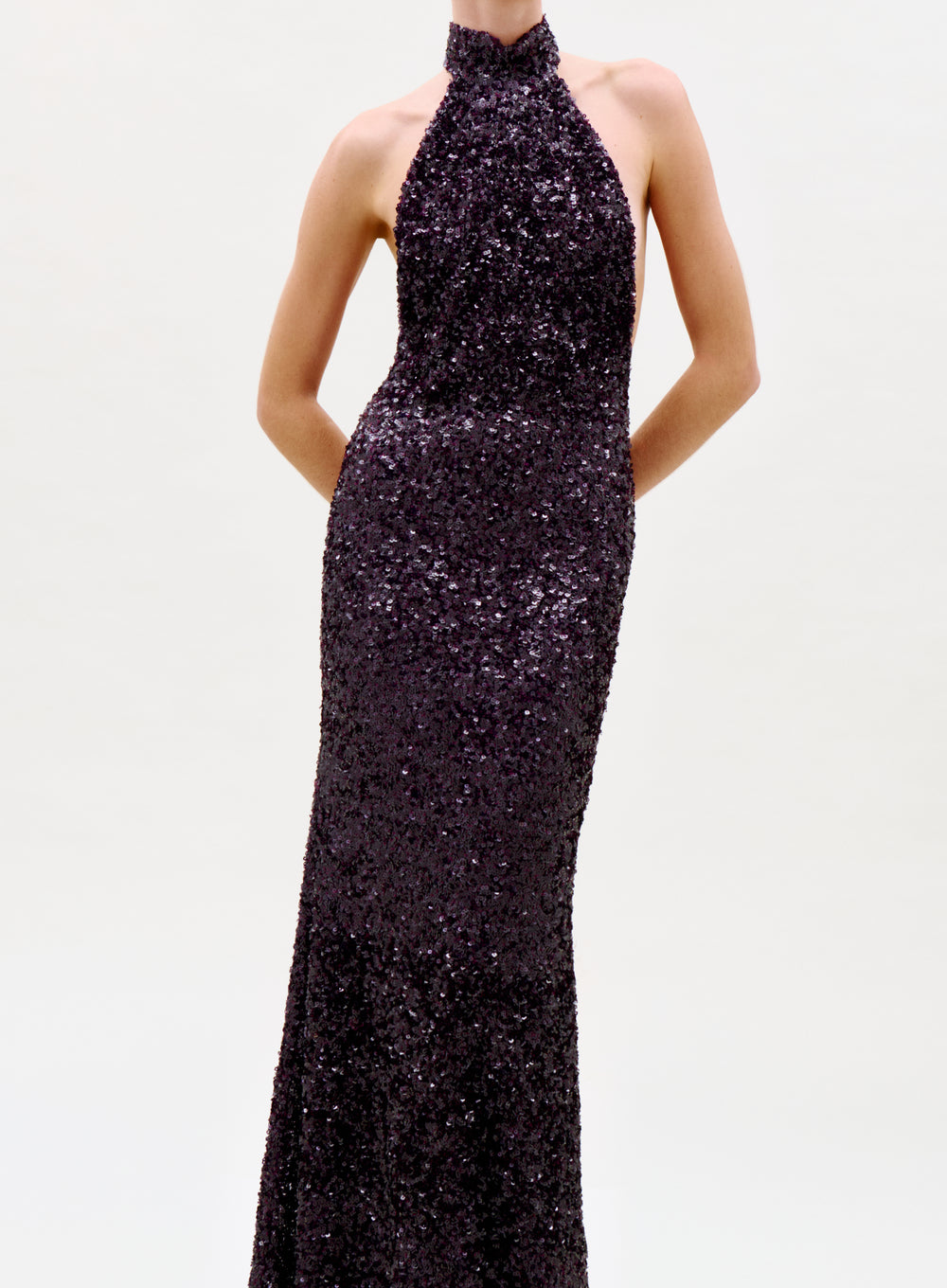 A person models the sleeveless, floor-length Dido Dress with dark sequins and a halter neck, standing hands behind their back against a plain light backdrop. The photo is cropped at the neck.