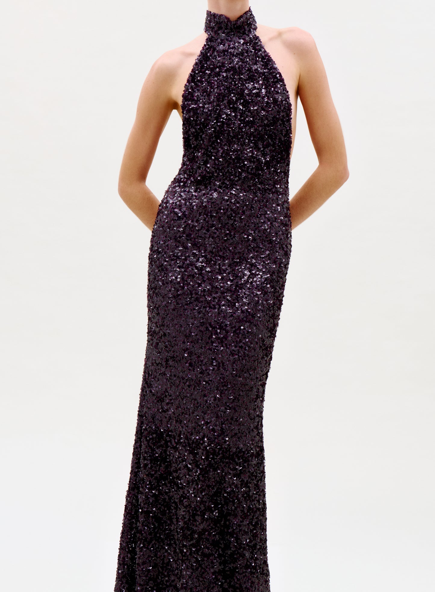 A person models the sleeveless, floor-length Dido Dress with dark sequins and a halter neck, standing hands behind their back against a plain light backdrop. The photo is cropped at the neck.