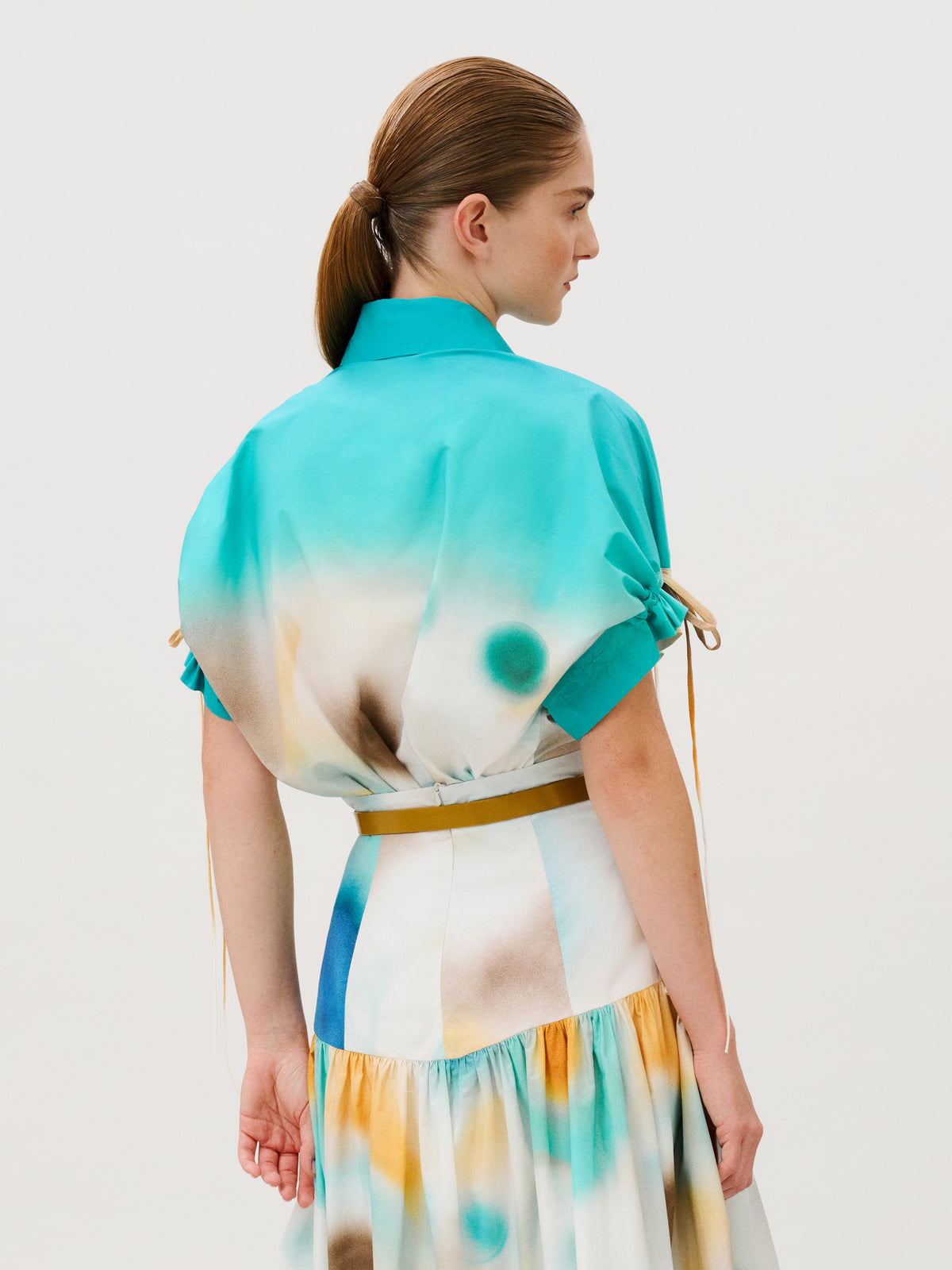 A person wearing a Dixie Shirt in a turquoise and white abstract print with drawstring sleeves and a thin tan belt.