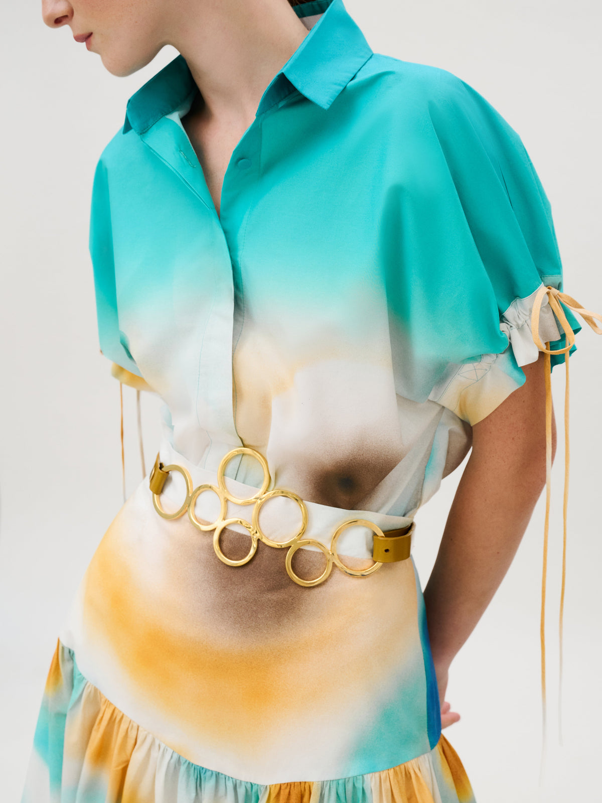 Dixie Shirt, a short-sleeved button-down crafted from Italian fabric with a teal, white, and yellow gradient print, featuring drawstring sleeves and a belt with interlocking gold circles.