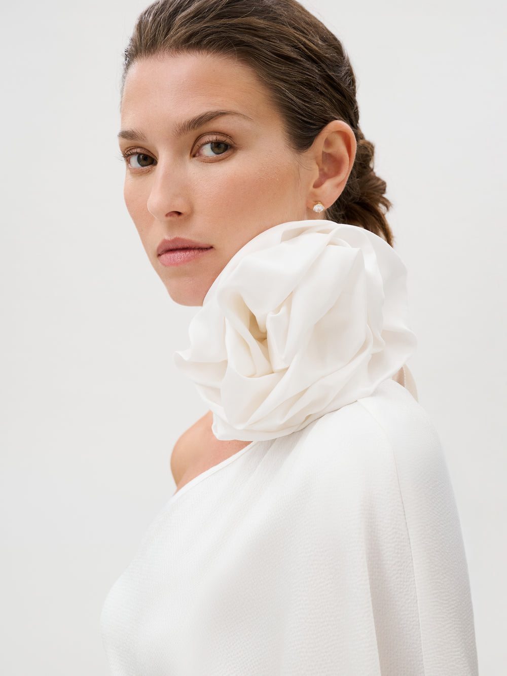 A woman wearing the Doli Dress—a white, off-shoulder piece featuring a large, ruffled collar—gazes confidently at the camera against a plain, light background. Her hair is pulled back to highlight the elegant garment, which is expertly crafted from hammered satin in Italy, adding an extra layer of sophistication to her poised presence.