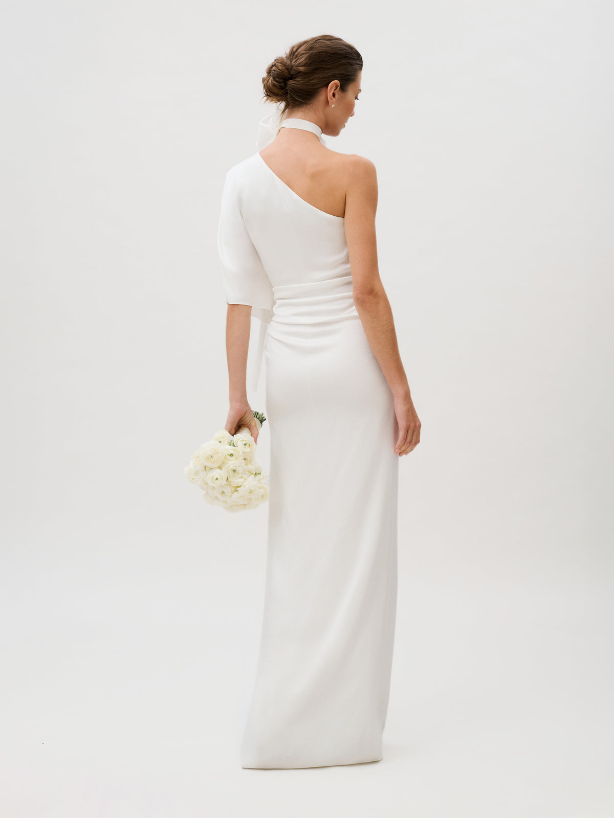 A woman stands with her back to the camera against a plain light background, holding a bouquet of white flowers and wearing the Doli Dress, a one-shoulder white hammered satin gown.