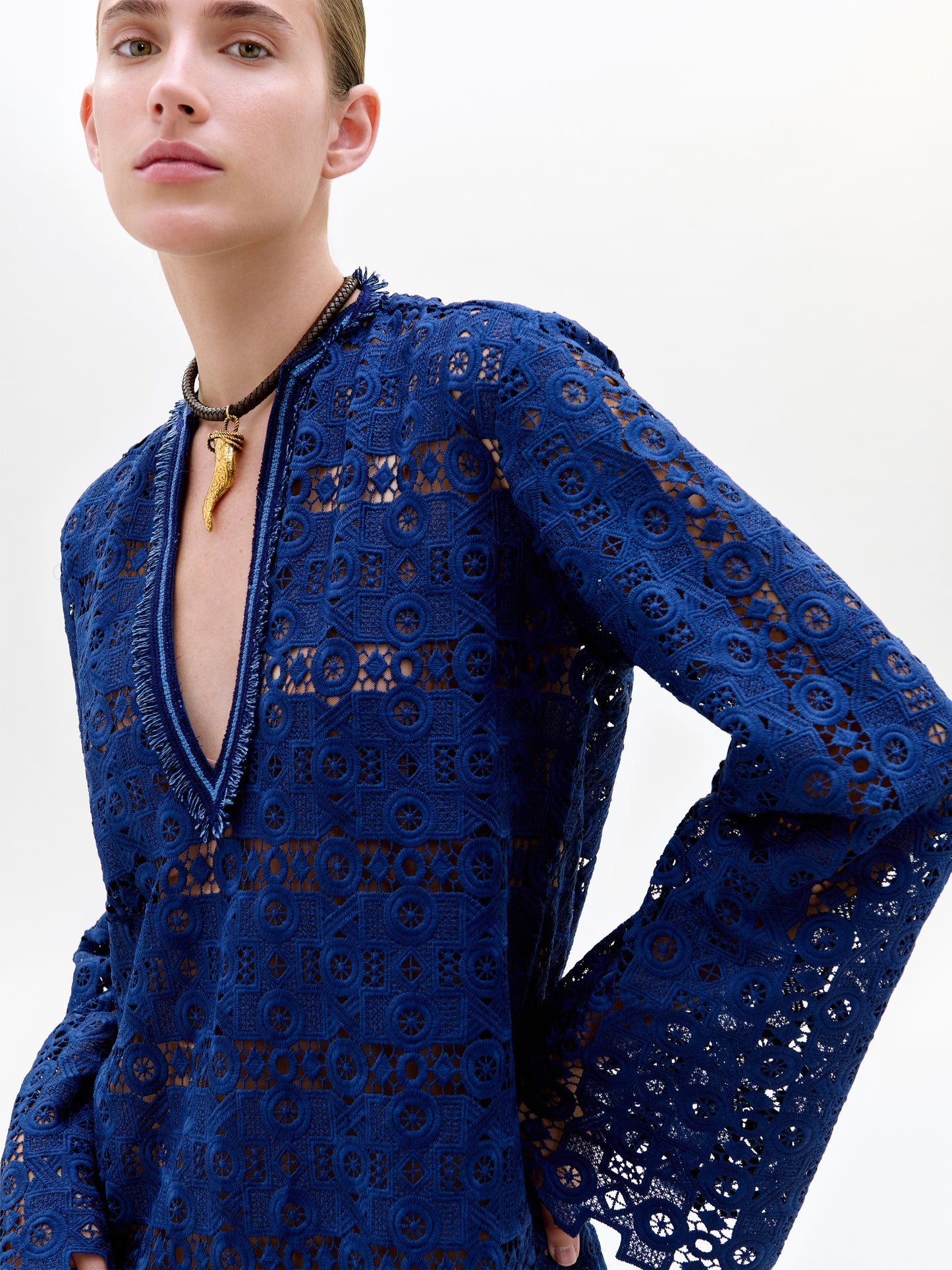 A person wearing the deep blue, long-sleeved Dorenia Dress made from Italian guipure lace with a deep V-neckline, paired with a black necklace with a gold pendant, stands against a plain white background.