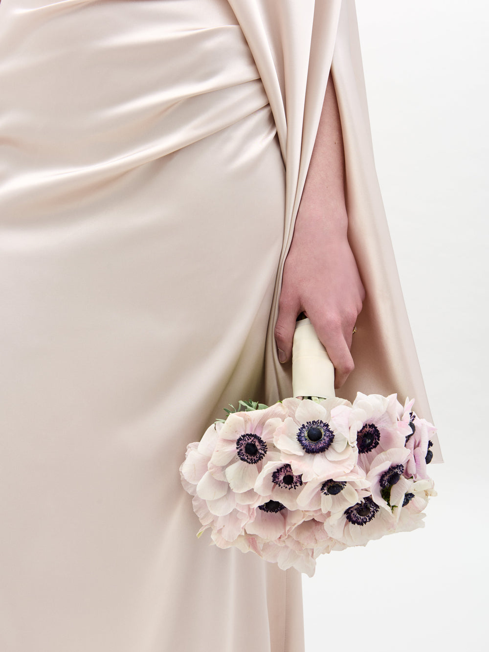 A person stands in a Dotty Dress, an off-the-shoulder satin gown, holding a bouquet of white and purple flowers against a plain white background.