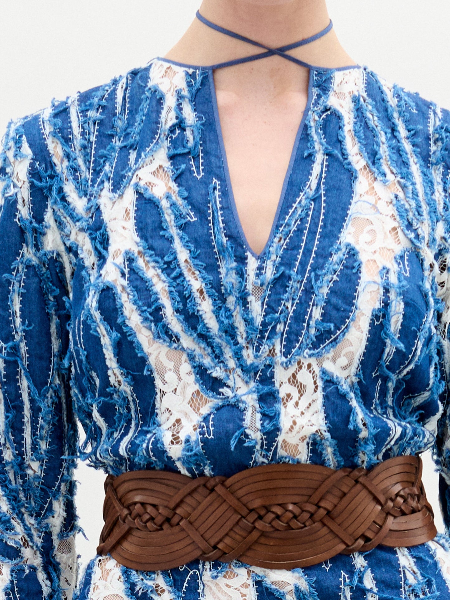 A person stands against a plain background wearing the Ebba Dress, a long blue and white patterned dress with a brown belt, high neckline, and subtle criss-cross detailing that enhances its elegant column silhouette.