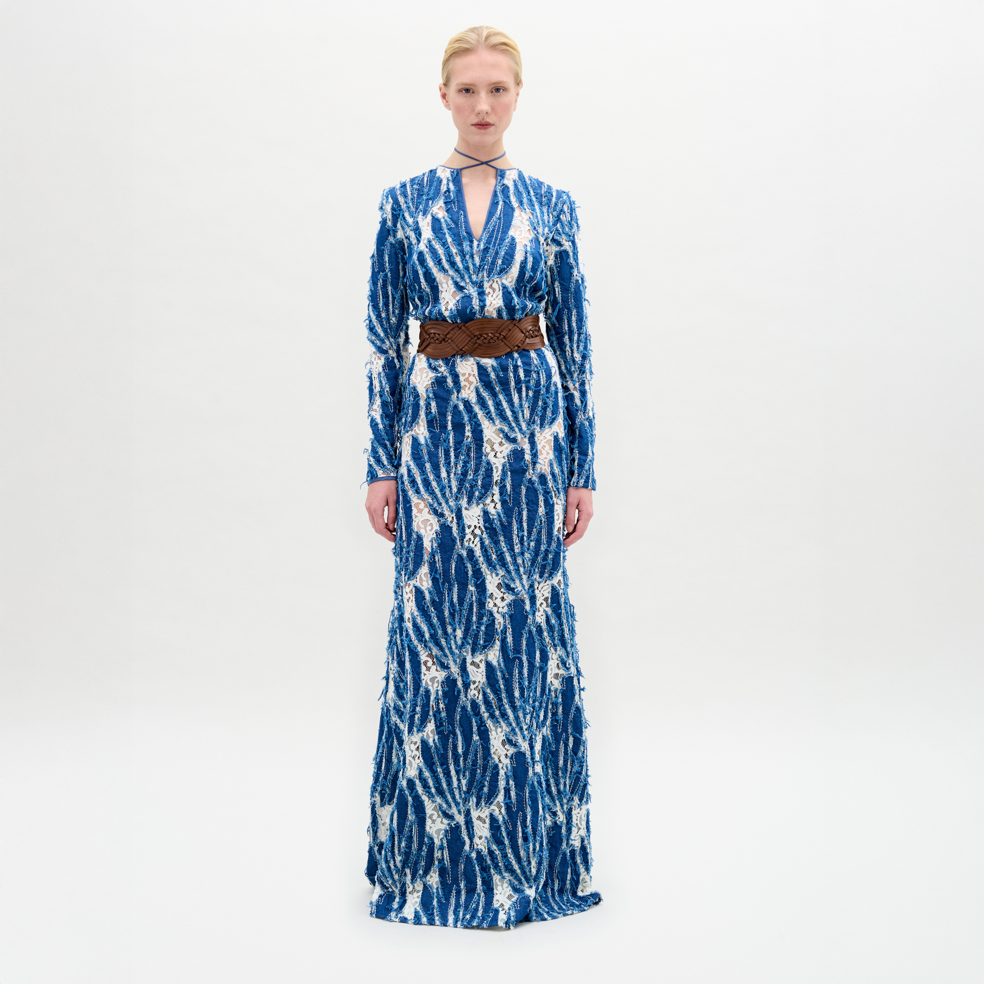 A person stands against a plain background wearing the Ebba Dress, a long blue and white patterned dress with a brown belt, high neckline, and subtle criss-cross detailing that enhances its elegant column silhouette.