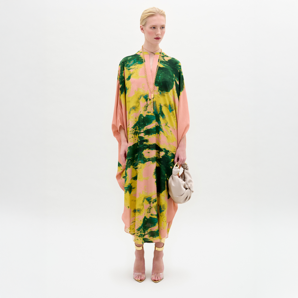 A woman stands against a white background wearing the Elea Tunic, an oversized 100% silk maxi dress with pink, yellow, and green patterns, paired with a beige bag and strappy heels.