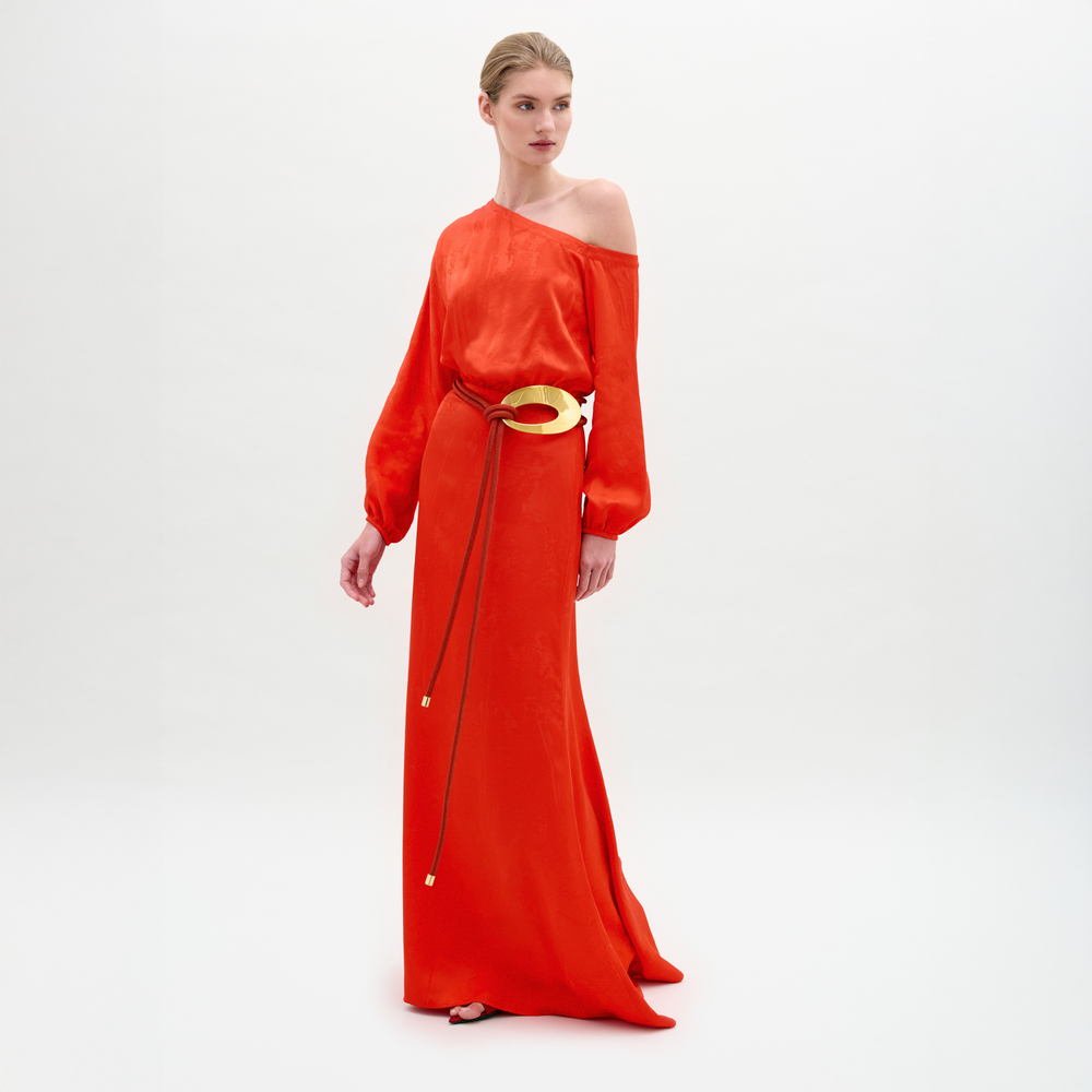 A woman poses against a white background in the Elga Dress—a red, floor-length, one-shoulder maxi dress with long sleeves and a gold 3D cord belt.