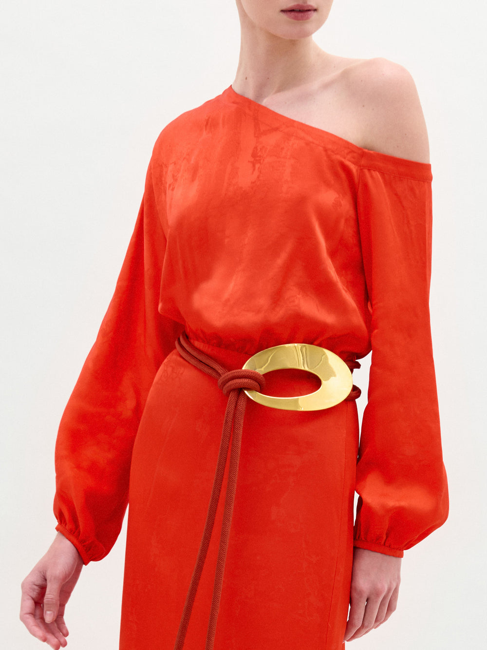 A woman stands against a plain white background wearing the Elga Dress, a bright orange Italian fabric one-shoulder maxi with long sleeves, a shiny gold statement belt, and brown rope detail.