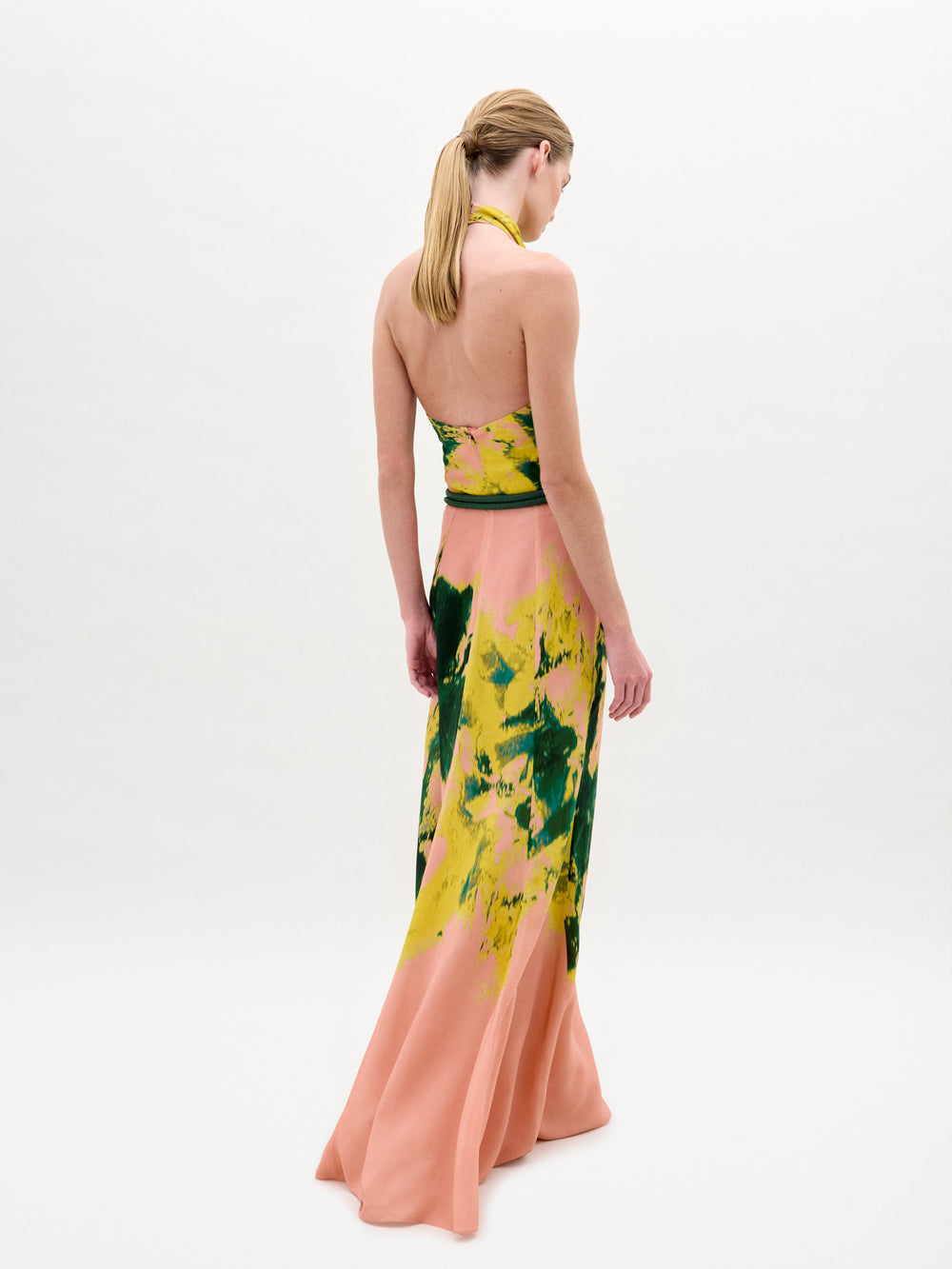 A woman wears the Ellis Dress—a sleeveless peach maxi with green and yellow abstract print, halter neckline, and side cutouts—against a plain white background.