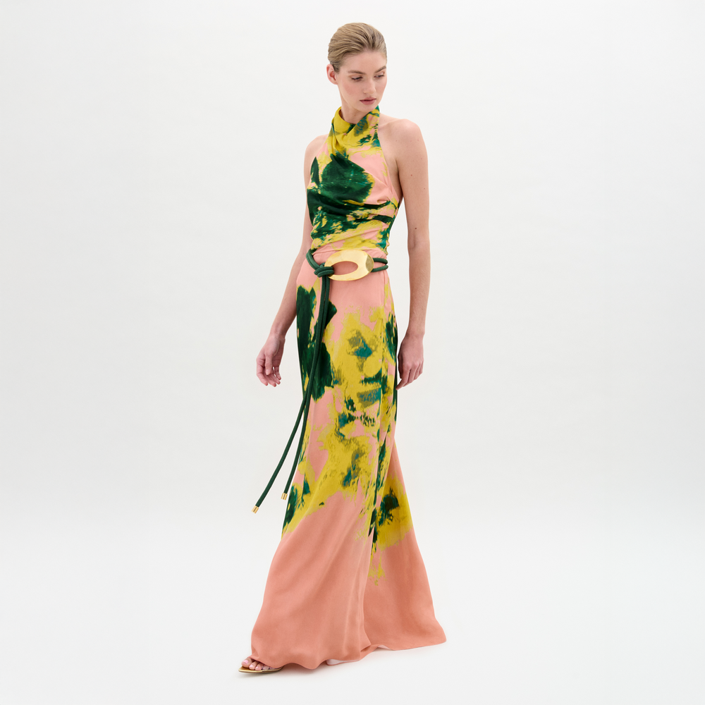 A woman wears the Ellis Dress—a sleeveless peach maxi with green and yellow abstract print, halter neckline, and side cutouts—against a plain white background.