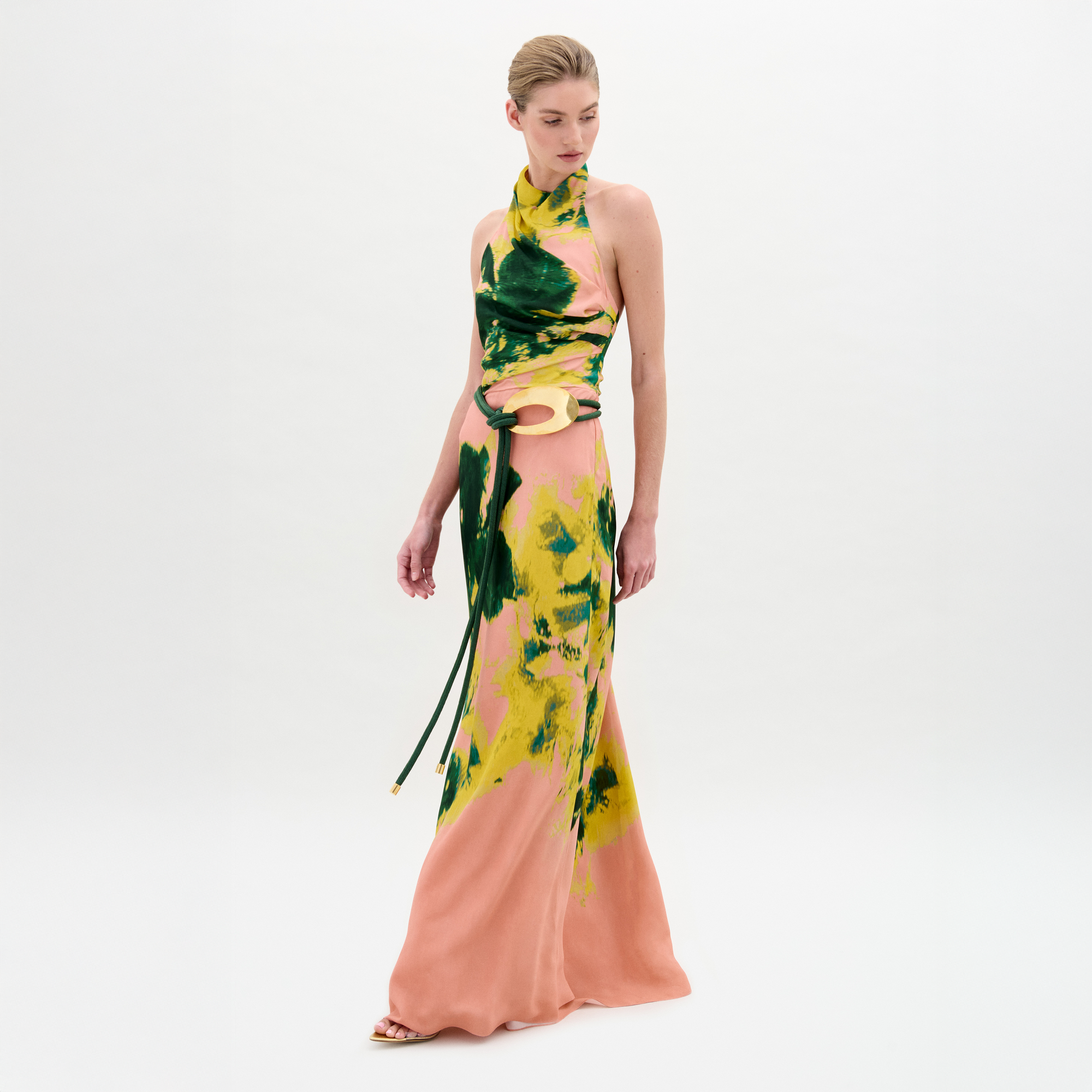 A woman wears the Ellis Dress—a sleeveless peach maxi with green and yellow abstract print, halter neckline, and side cutouts—against a plain white background.