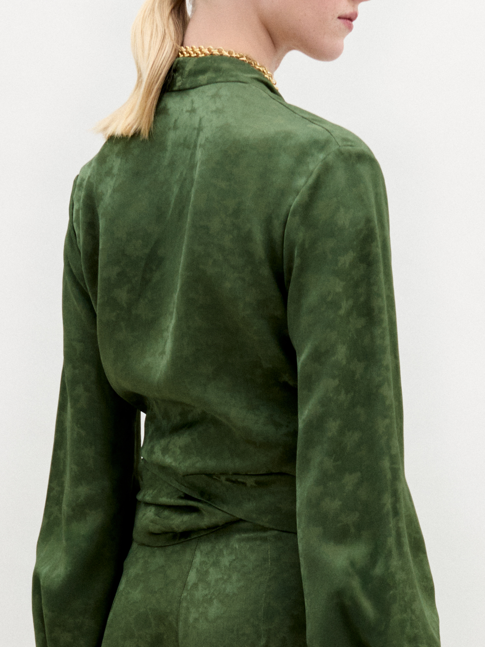 Person standing against a plain background wearing the green Elvie Blouse, a long-sleeved wrap top with wrap-around ties, matching pants, accessorized with a gold necklace and holding a gold handbag.